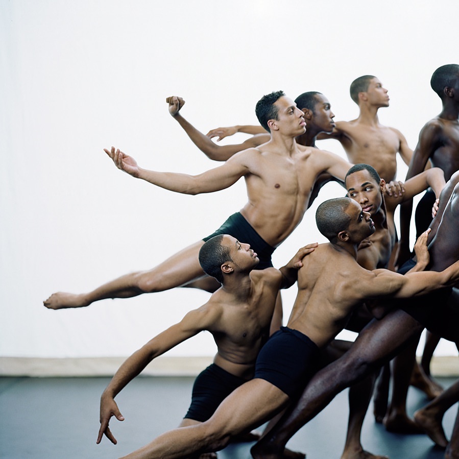 A group of athletic, bare-chested male dancers in motion performing choreography in a bright studio with white walls.