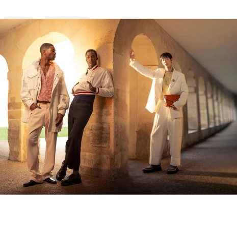 Three stylish adult male models pose in a covered walkway with arches, wearing fashionable outfits with jackets and trousers.