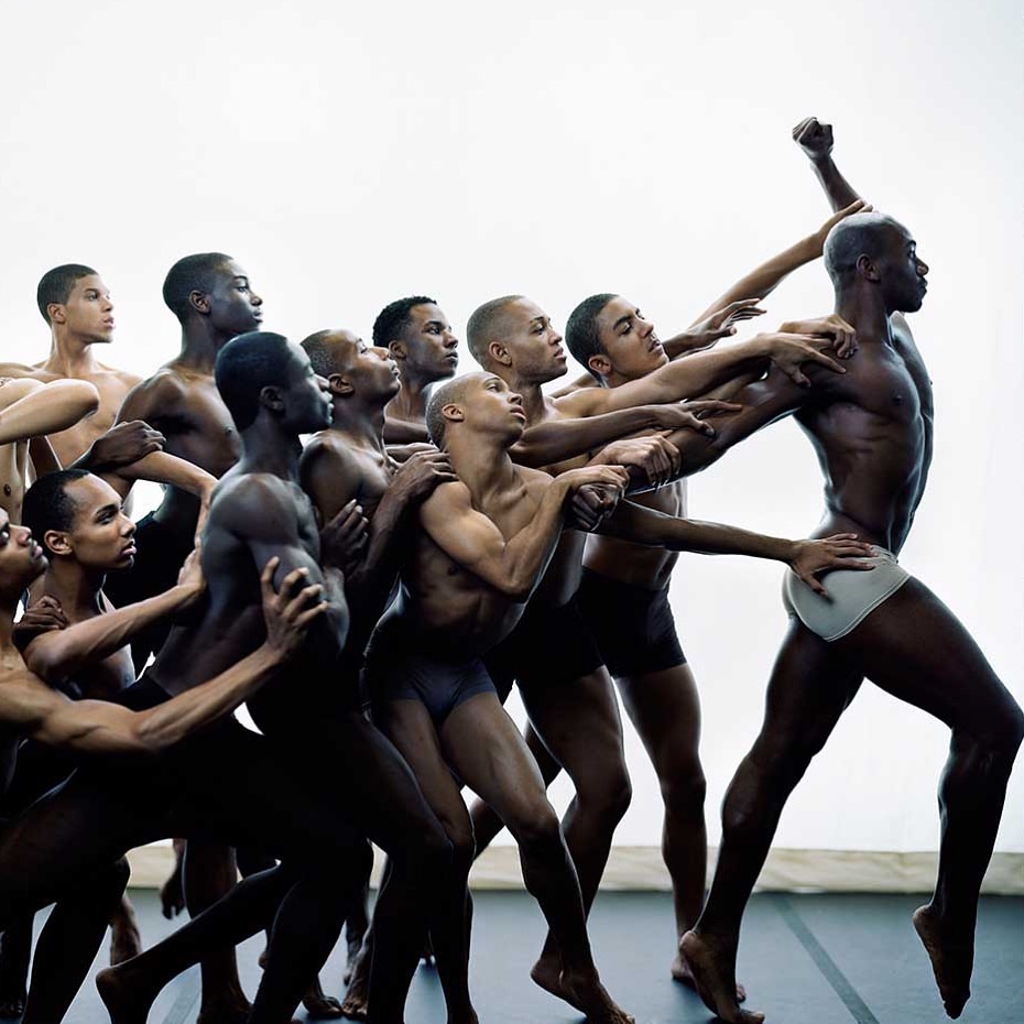 Group of male dancers in a performance line, with dark skin tones and sleeked hair, wearing minimal clothing, set against a plain white backdrop.