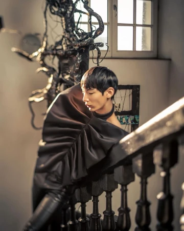 A man in a sculptural dark garment leans against a wooden staircase railing indoors in Paris.