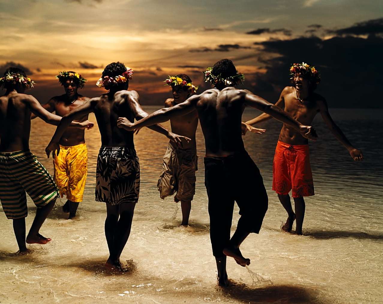 Group of men in swimwear and flower crowns stand in shallow ocean water at sunset, displaying traditional island culture dance.