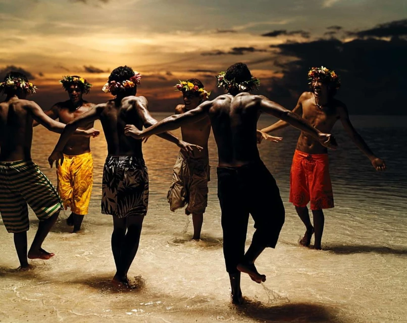 Group of men in swimwear and flower crowns stand in shallow ocean water at sunset, displaying traditional island culture dance.