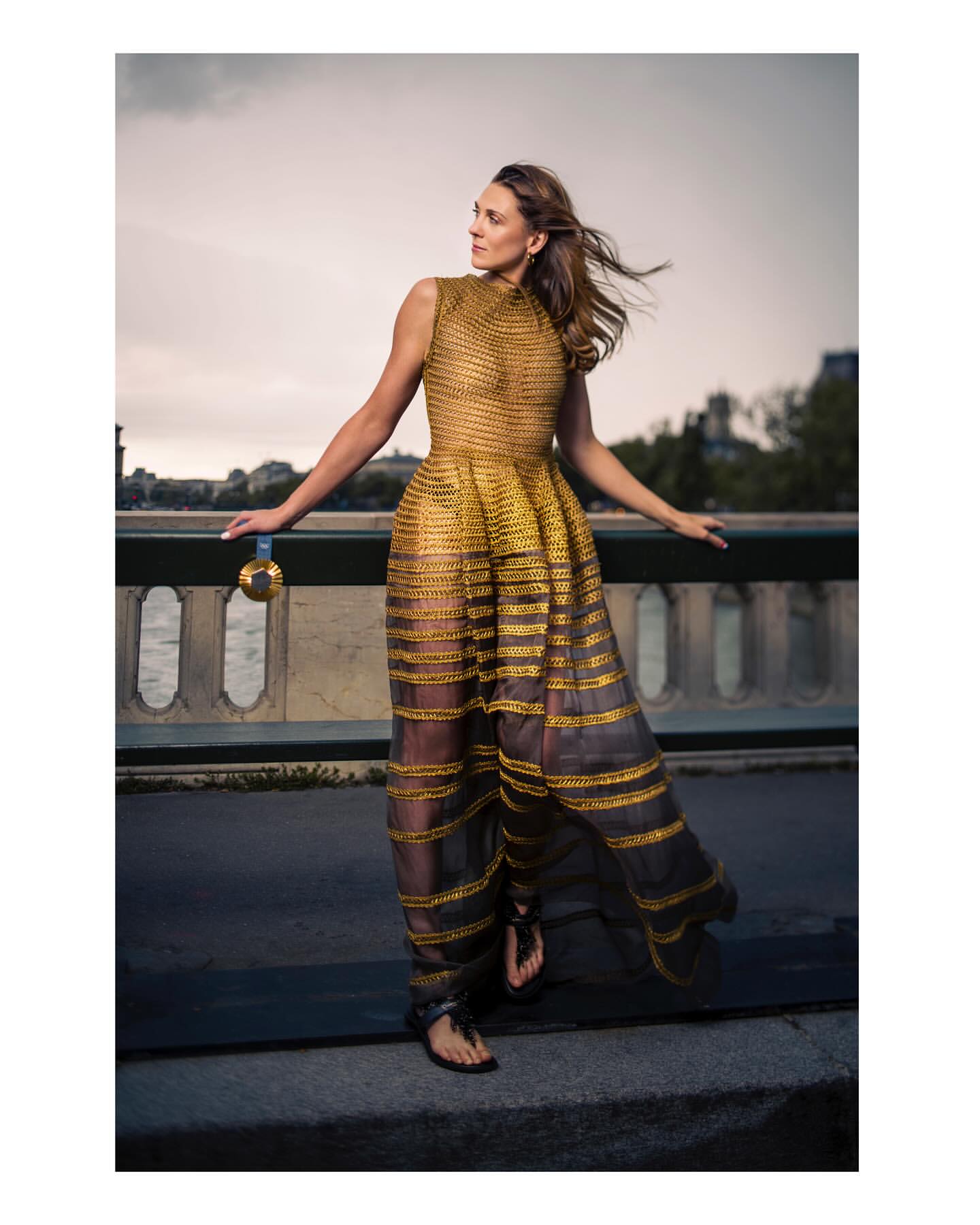 Woman in flowing gold dress stands on a bridge overlooking a river in an outdoor fashion shot.