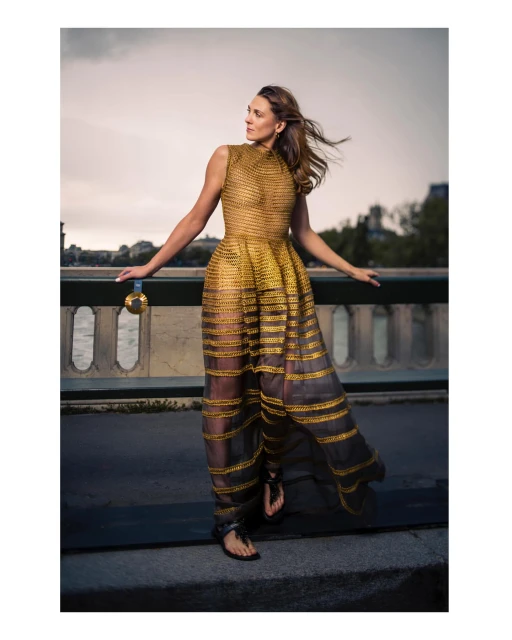 Woman in flowing gold dress stands on a bridge overlooking a river in an outdoor fashion shot.