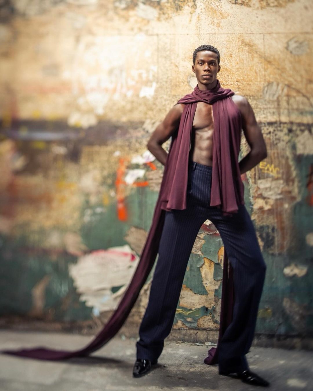 Stylish male model poses in draped fabric and pinstripe pants against a weathered urban wall, highlighted by studio lighting and shadow.