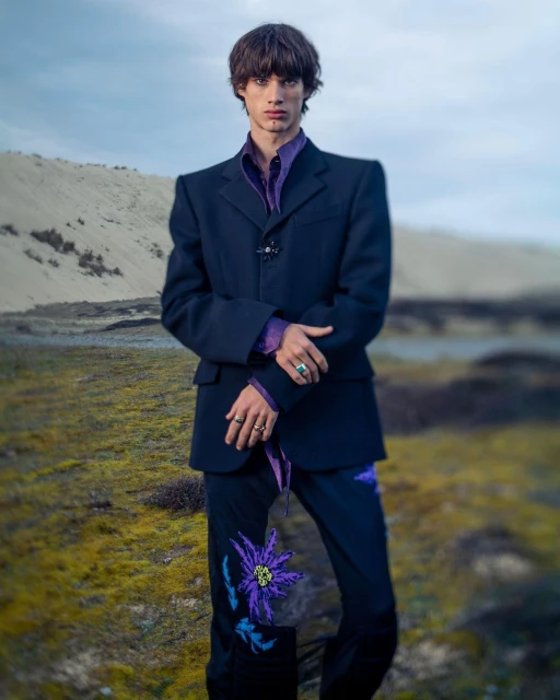 An adult male fashion model poses in an outdoor environment wearing a dark blazer and pants with flower embroidery, plus a purple collared shirt.