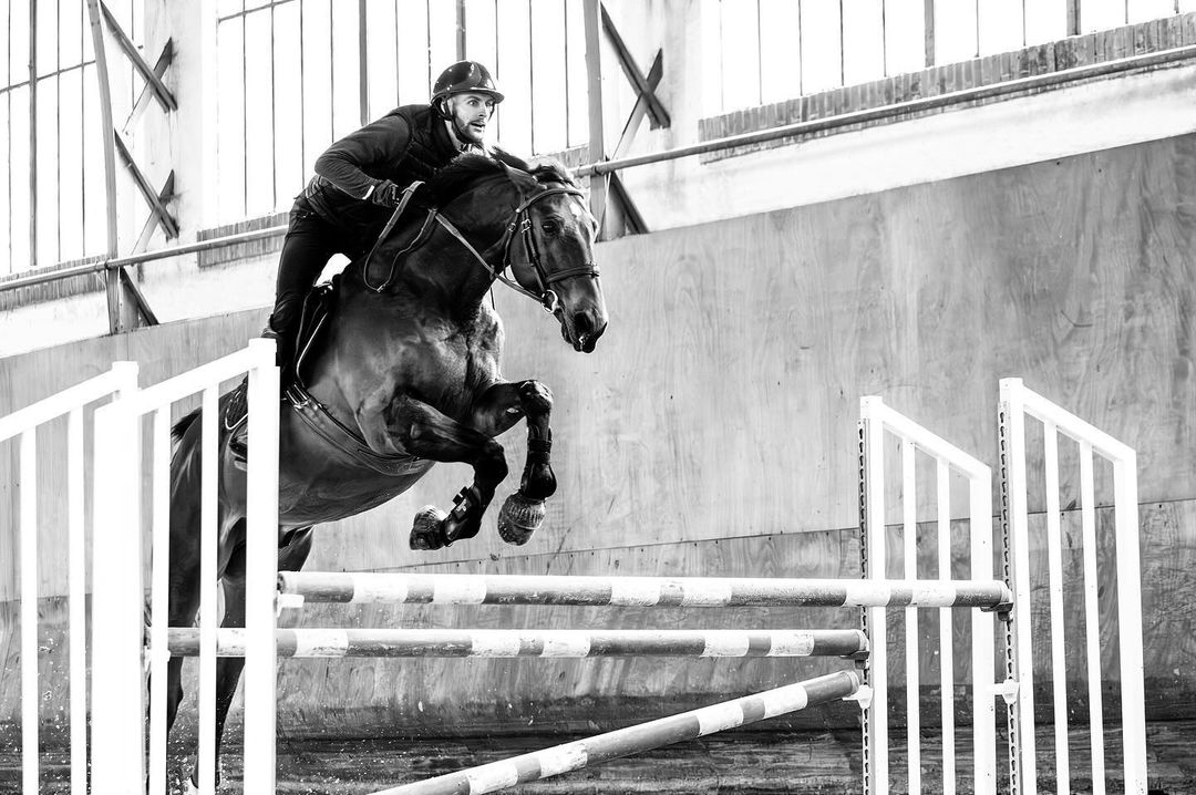 Equestrian action: a man in helmet rides a dark horse jumping over a white and red striped fence inside an arena with bright lighting.