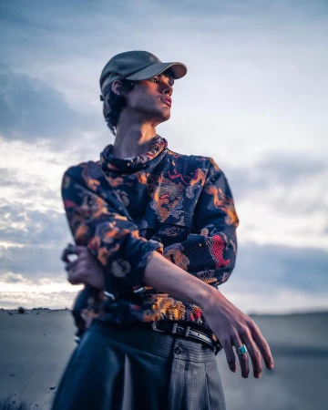 Young male model wearing patterned shirt and baseball cap posing outdoors against an overcast sky