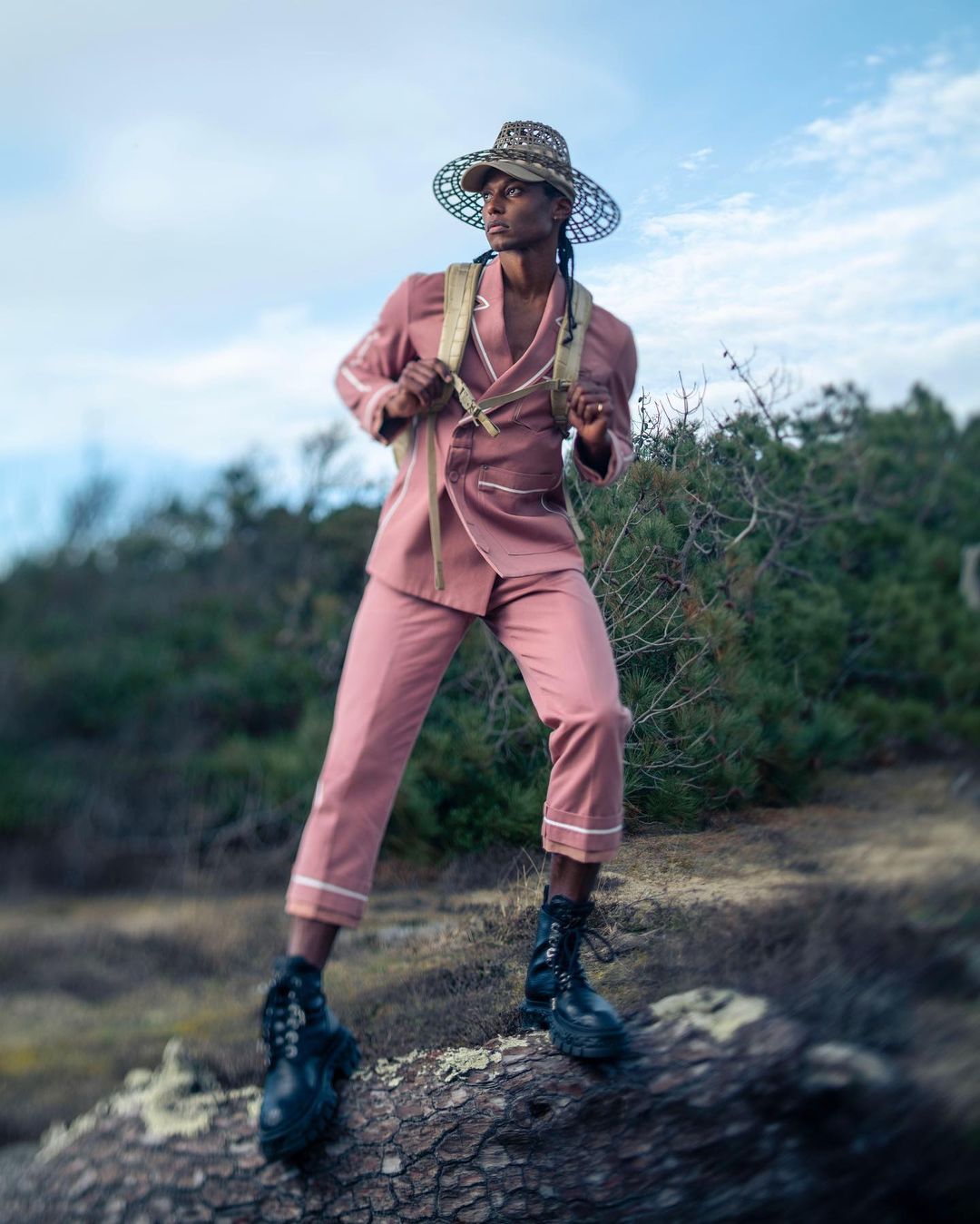 A person in a pink suit and decorative hat standing on a log outdoors against a sky and foliage background for a fashion shoot.