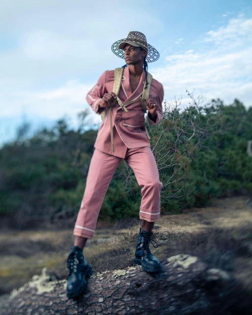 A person in a pink suit and decorative hat standing on a log outdoors against a sky and foliage background for a fashion shoot.