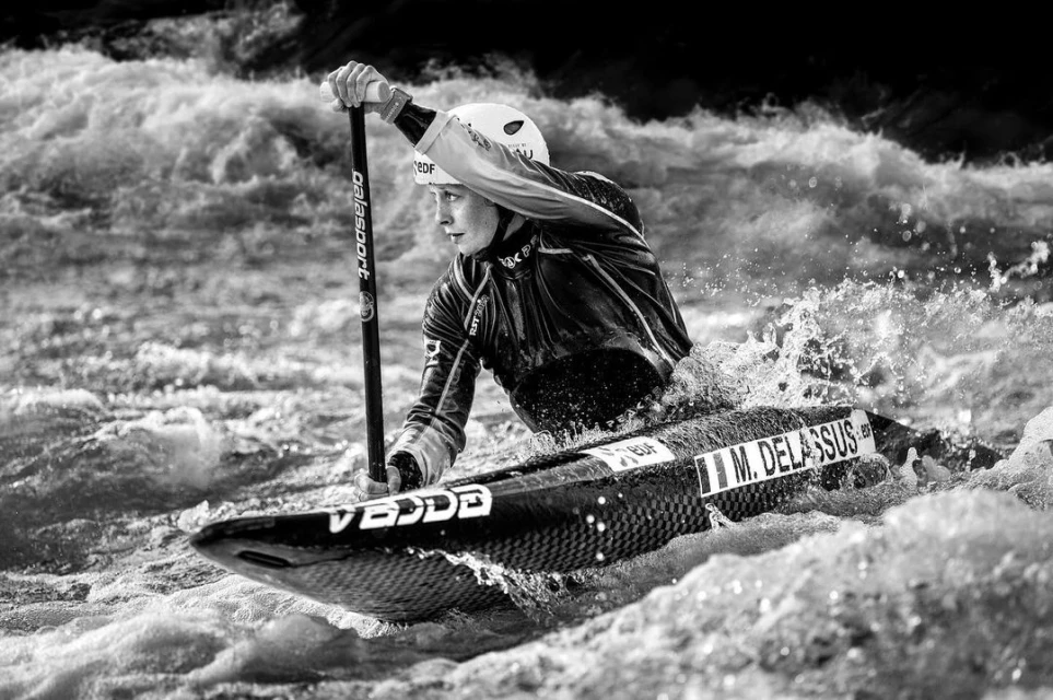 A woman paddles a kayak through whitewater rapids, holding an oar, wearing a helmet and water gear in a black and white action shot.