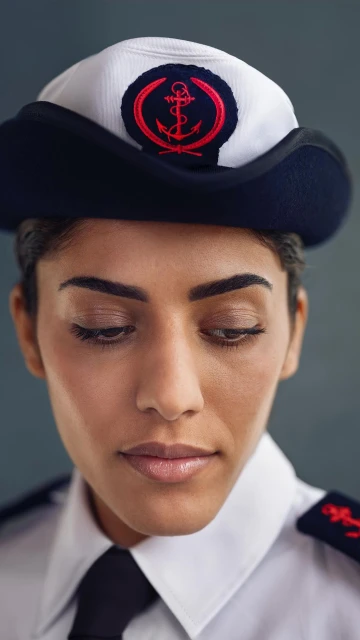 Close-up of a woman with tan skin wearing a navy and white sailor hat with an anchor, a white shirt, and a black tie.