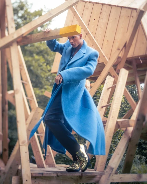 Stylish man poses in a blue coat and gold boots within a unique wooden structure outdoors, surrounded by lush green foliage.