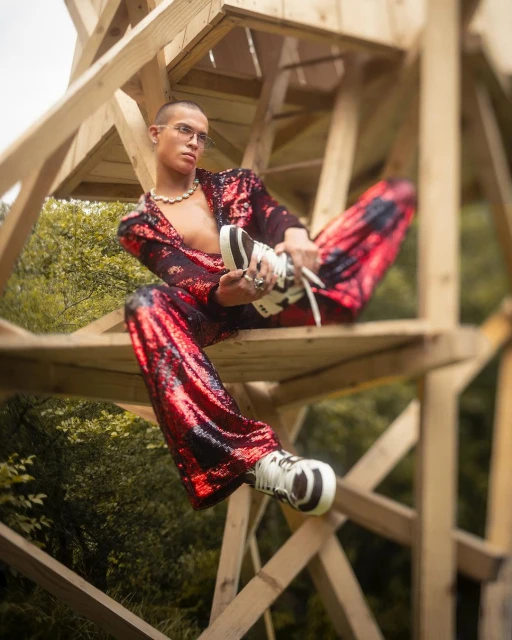 Stylish adult male poses in red sequin suit and sneakers on a wooden structure in nature. Fashion editorial outdoor shot.