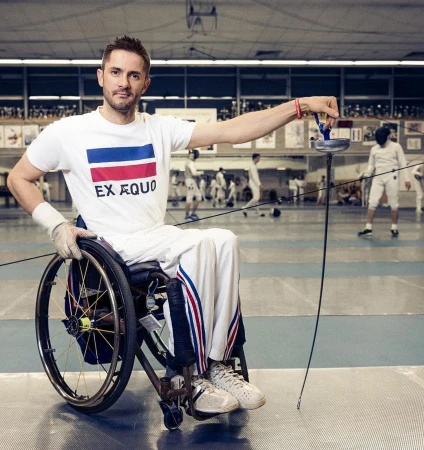 Adaptive athlete in wheelchair fencer attire holds a foil in a fencing hall, competing in the inclusive sport of wheelchair fencing.