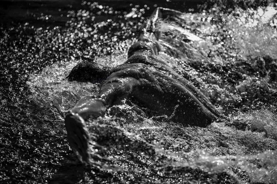 Black and white photo of an adult swimming in water, the swimmer's body and arms are visible, splashing and rippling the water.