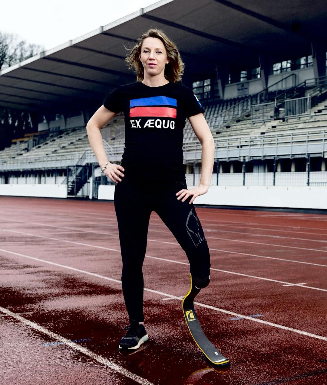 A woman with a prosthetic leg in athletic wear poses on a wet track, an athletic stadium visible in the background.
