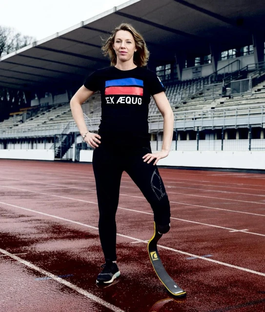 A woman with a prosthetic leg in athletic wear poses on a wet track, an athletic stadium visible in the background.