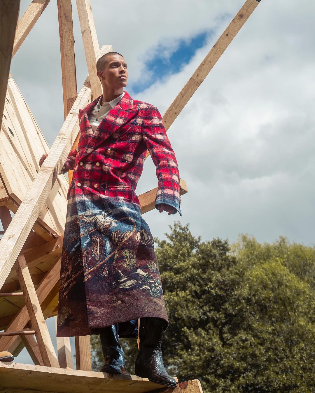 Man wearing a long red plaid coat with a landscape motif standing amidst raw wooden framing against a sky background.