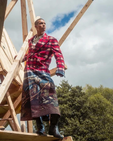 Man wearing a long red plaid coat with a landscape motif standing amidst raw wooden framing against a sky background.