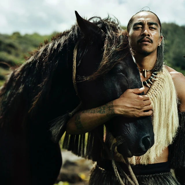Portrait of a Polynesian man embracing a black horse outdoors with a background of green hills and cloudy sky, showcasing cultural attire and connection.