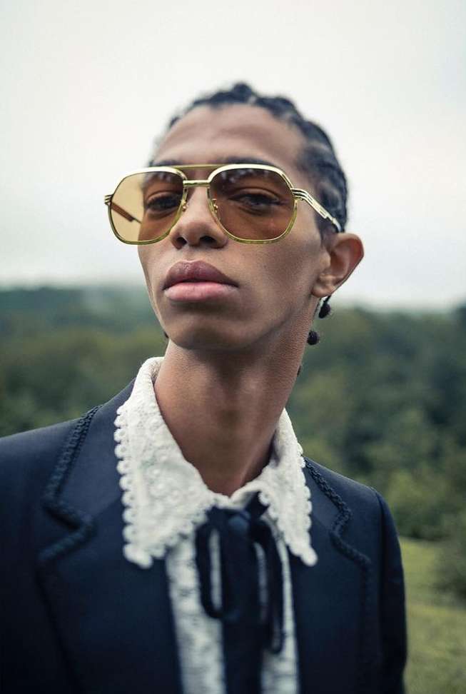 Close-up of a person wearing gold-rimmed sunglasses, a ruffled lace collar, and a dark suit against a blurred natural background.