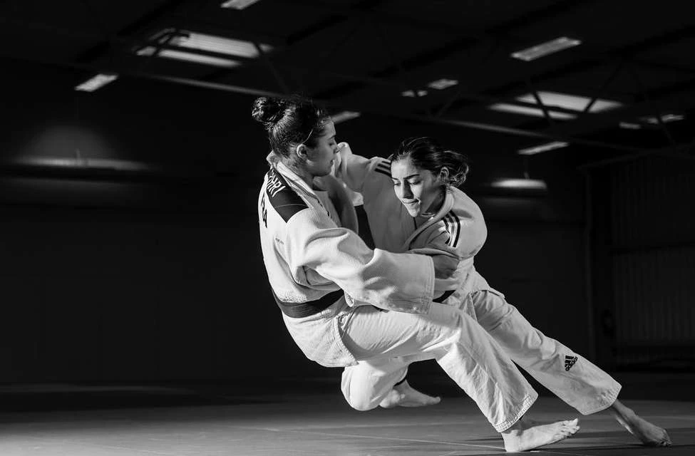 Two women judoka in a black and white action shot grappling on a mat in a training facility, focused on their competitive interaction.