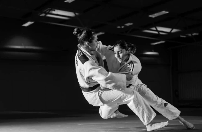 Two women judoka in a black and white action shot grappling on a mat in a training facility, focused on their competitive interaction.