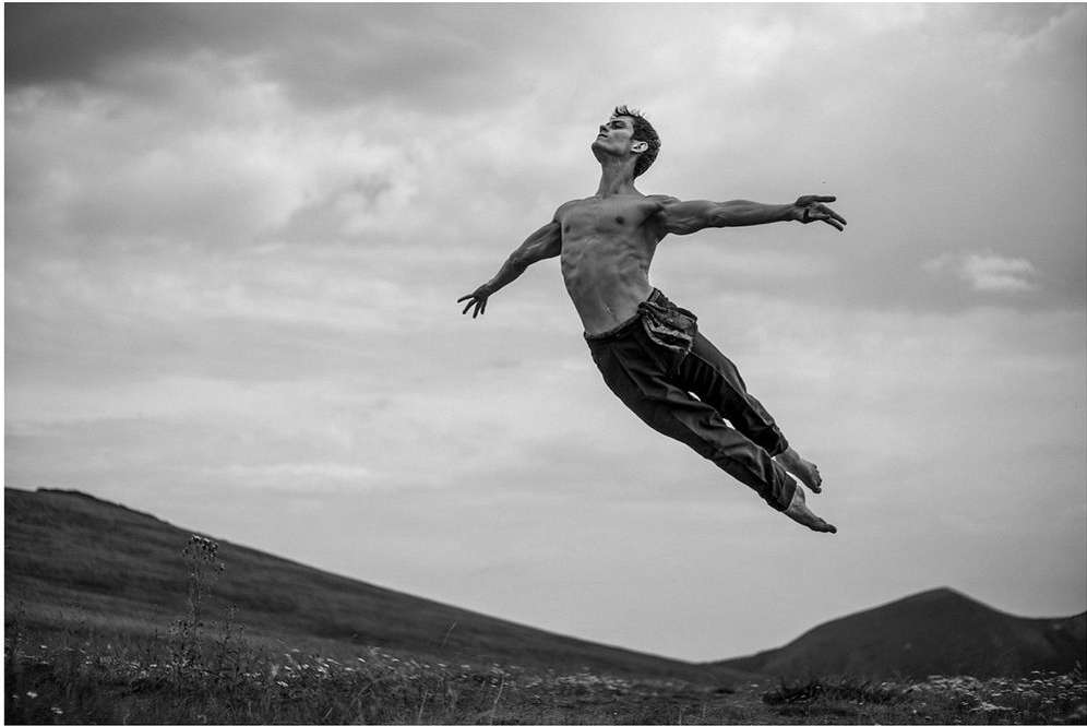 An adult male is captured mid-air in a dynamic pose against a backdrop of mountains and cloudy sky, creating a striking black-and-white image.
