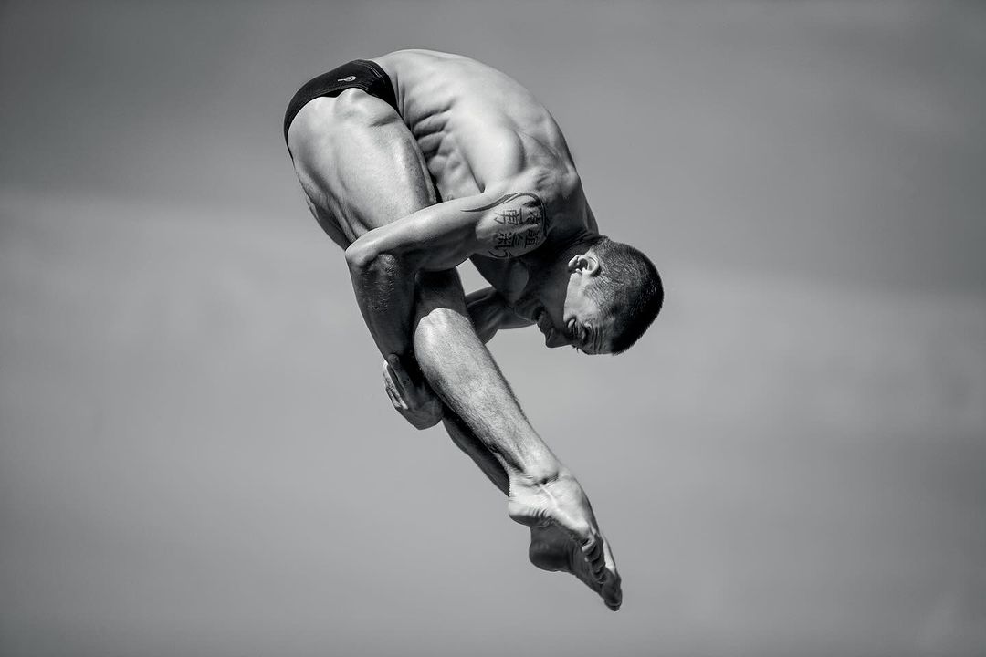 Monochrome full shot of a muscular adult male athlete diving in a curled position with a grey sky background.