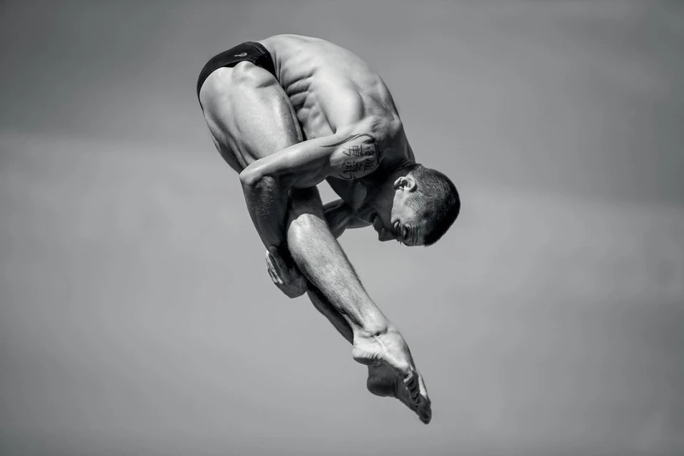 Monochrome full shot of a muscular adult male athlete diving in a curled position with a grey sky background.