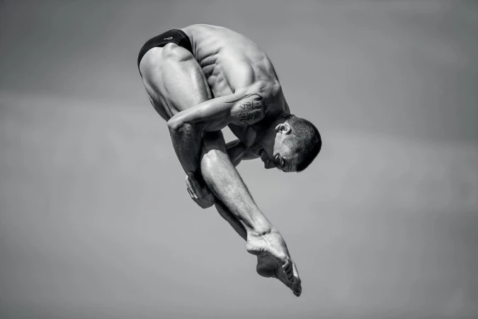 Monochrome full shot of a muscular adult male athlete diving in a curled position with a grey sky background.