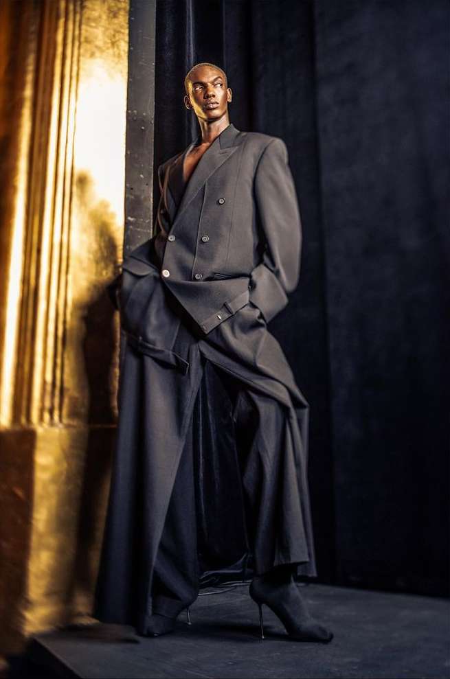 A slender Black male model in a draped black suit and heeled boots poses against gold columns, conveying high fashion in a studio setting.
