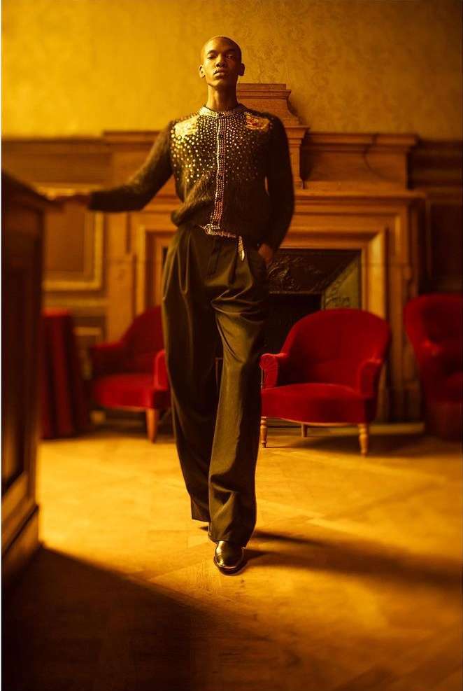 A bald, dark-skinned man in a patterned black jacket and black pants is posing in an indoor space with red chairs.
