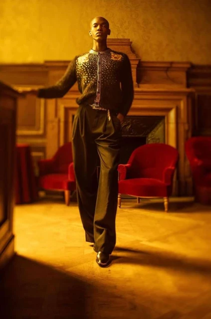 Full-shot of a Black male model wearing an embellished cardigan and wide leg pants standing indoors with decorative fireplace.