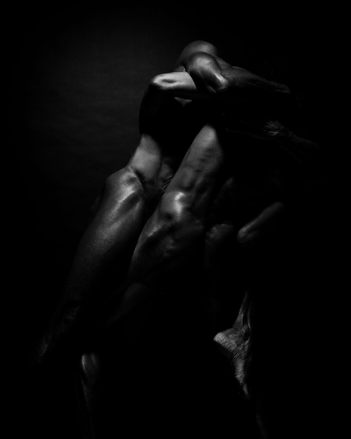 Close-up monochromatic black and white photograph of intertwined muscled human bodies forming an abstract sculptural shape in deep shadow.