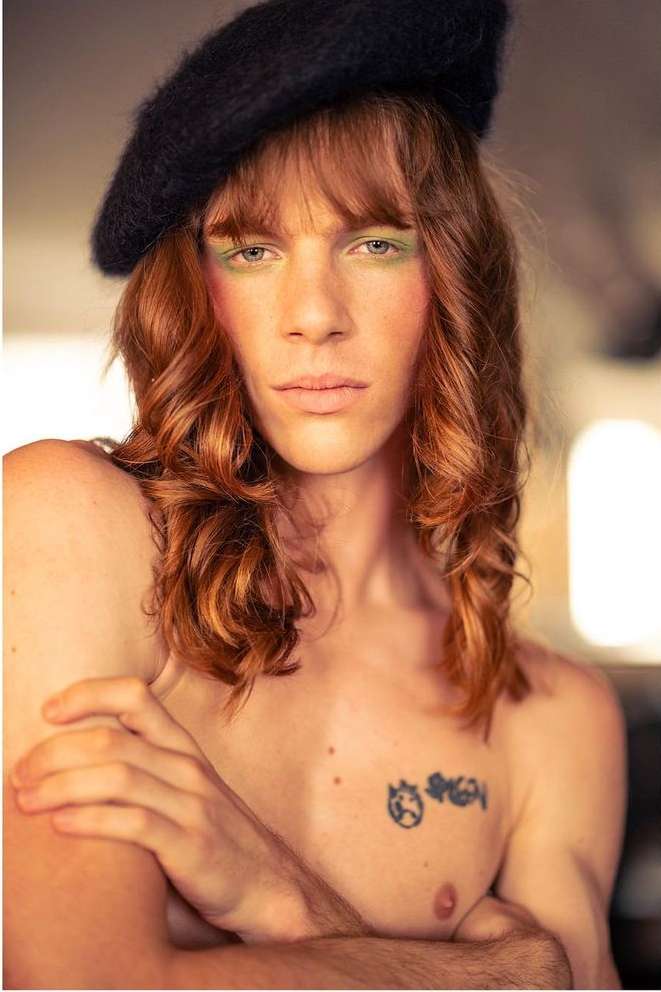 Portrait of adult male with red wavy hair wearing a black beret, green eyeshadow, with tattoo on his chest in soft light.