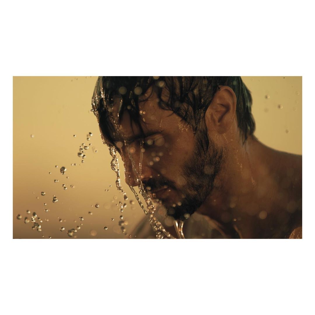 Adult male with a beard stands with his head tilted back, getting wet with water in a golden tinted indoor shower or washroom.