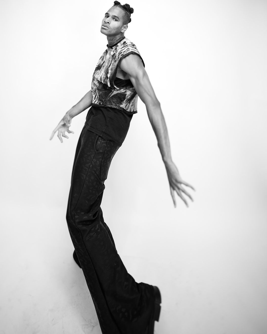 Black and white portrait of an adult with dark hair styled in buns, wearing a crop top and wide leg pants against a plain background.