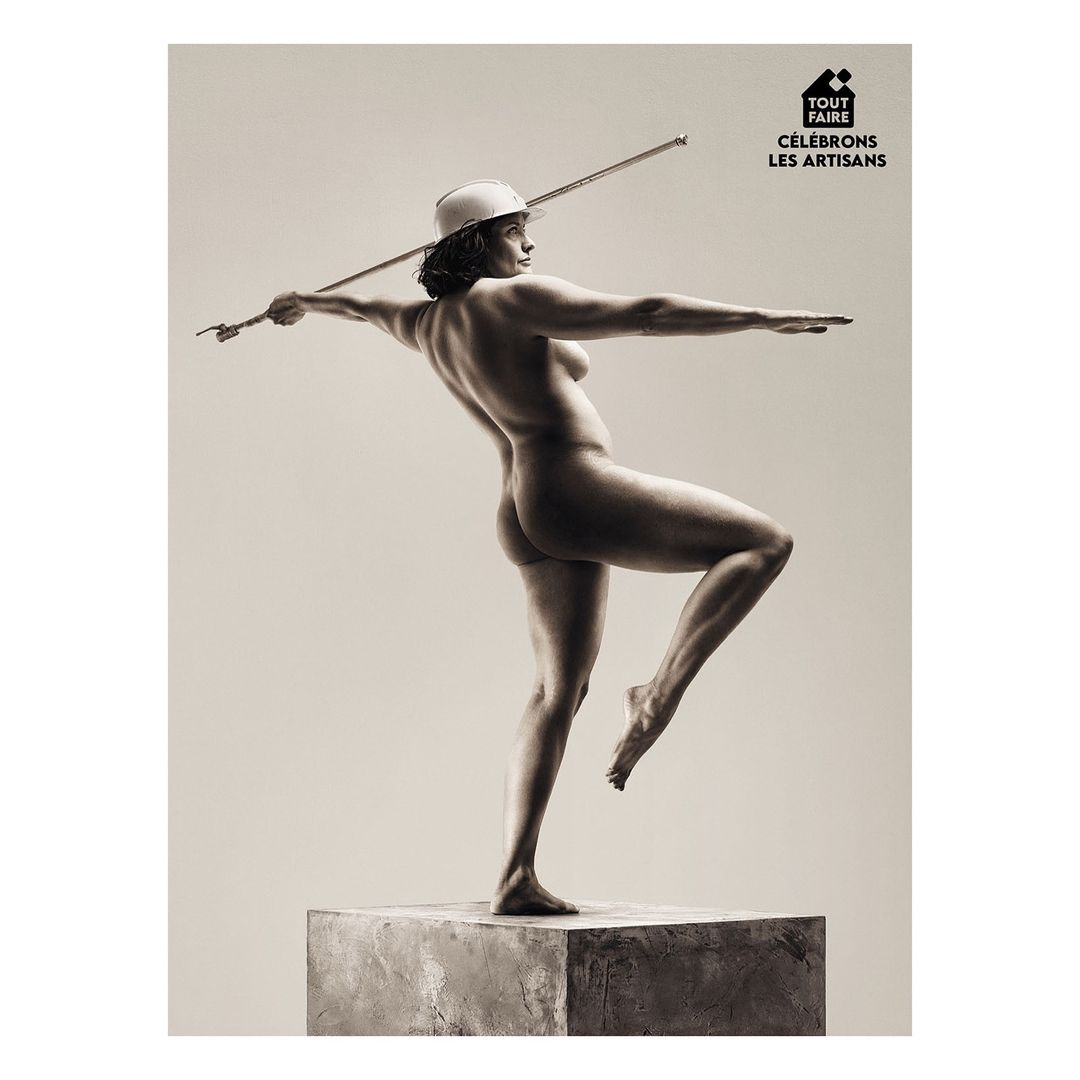 Sepia-toned art photo of a nude adult female balancing on a concrete block, wearing a construction hat, and holding a metal rod.