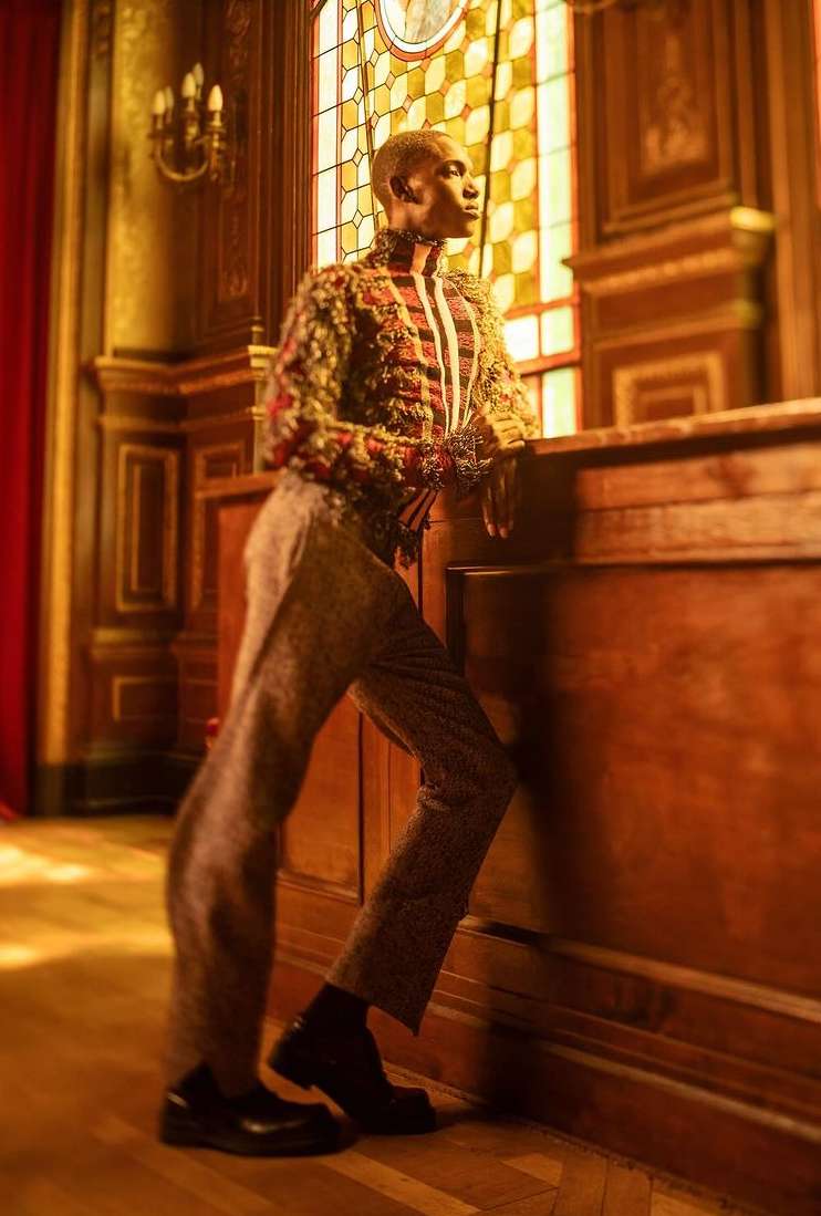 Full-length portrait of a person wearing a patterned jacket and trousers, leaning against a wooden panel near a stained glass window.