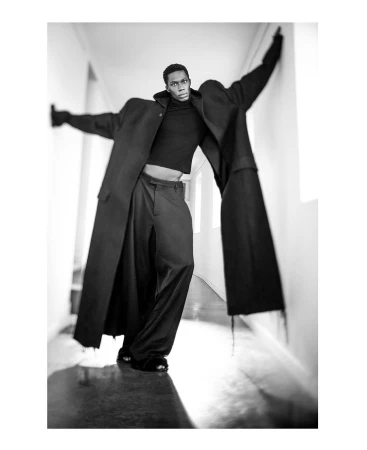 Black man wearing an oversized black coat and trousers leaning in a narrow hallway