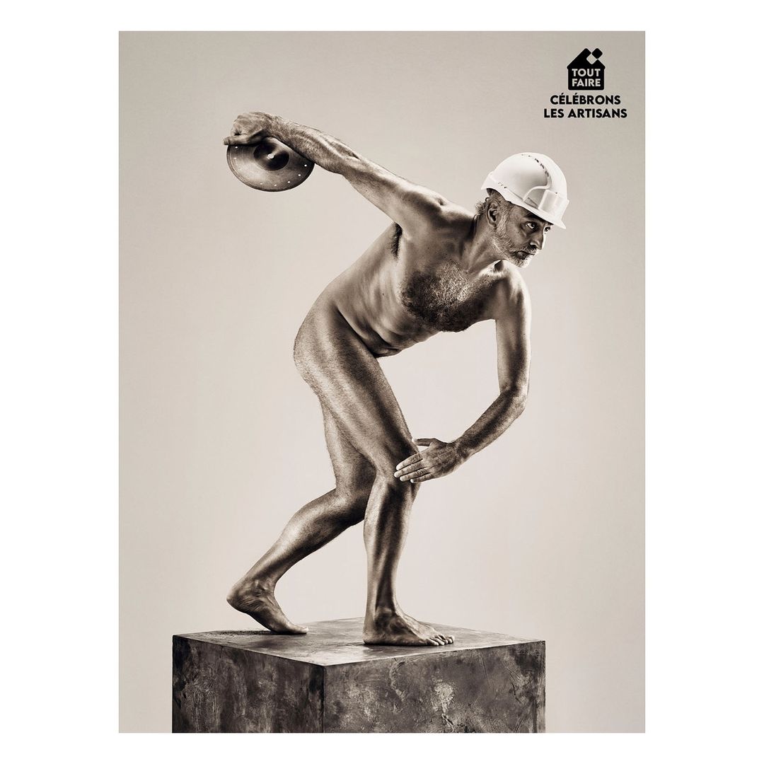 Artisan in nude pose throwing discus while wearing hard hat stands on pedestal against light grey backdrop in studio setting.
