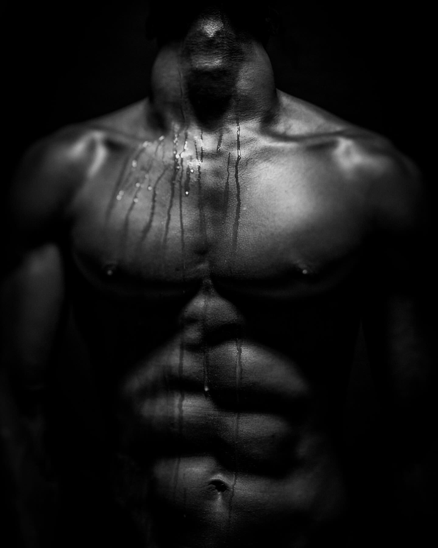 Monochrome studio shot of a muscular, partially visible adult male torso with water droplets, set against a dark background.