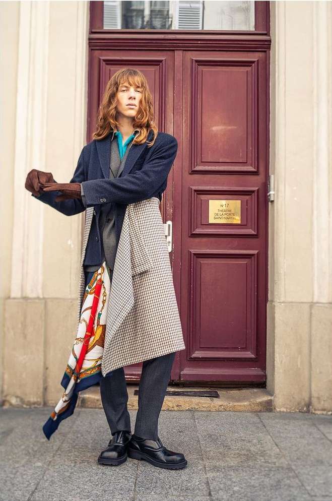 Red-haired adult poses in a navy jacket, grey vest, and patterned scarf in front of a maroon door and cream facade in urban setting.