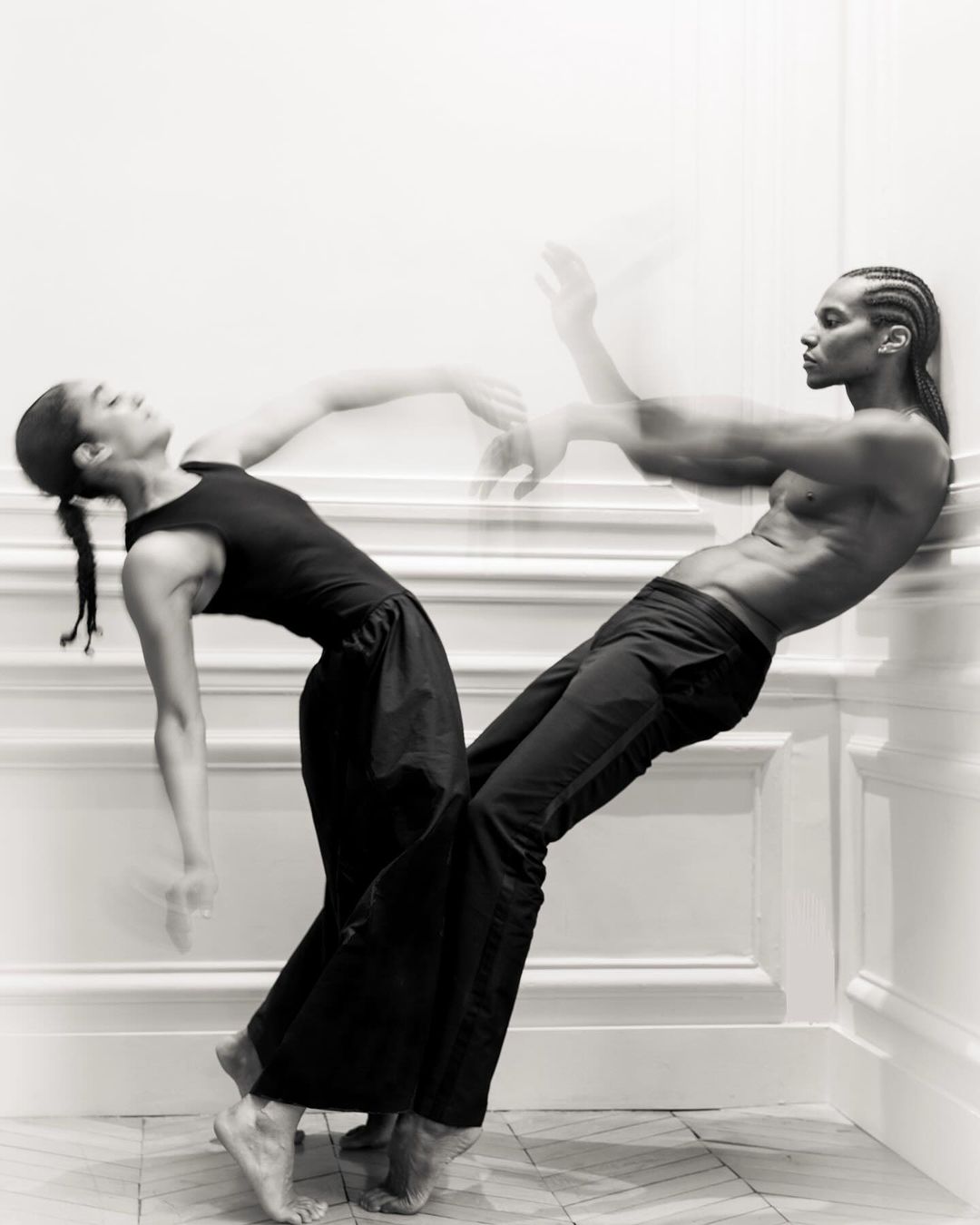 Two dancers, an adult woman and a shirtless adult male, lean against a white wall on a herringbone tile floor in a black and white photo.