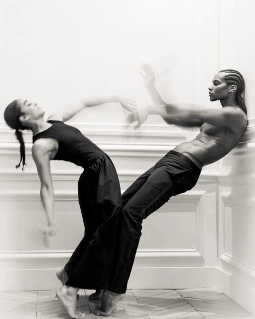 Two dancers, an adult woman and a shirtless adult male, lean against a white wall on a herringbone tile floor in a black and white photo.
