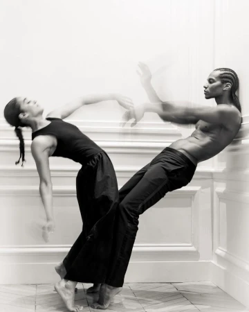 Two dancers, an adult woman and a shirtless adult male, lean against a white wall on a herringbone tile floor in a black and white photo.