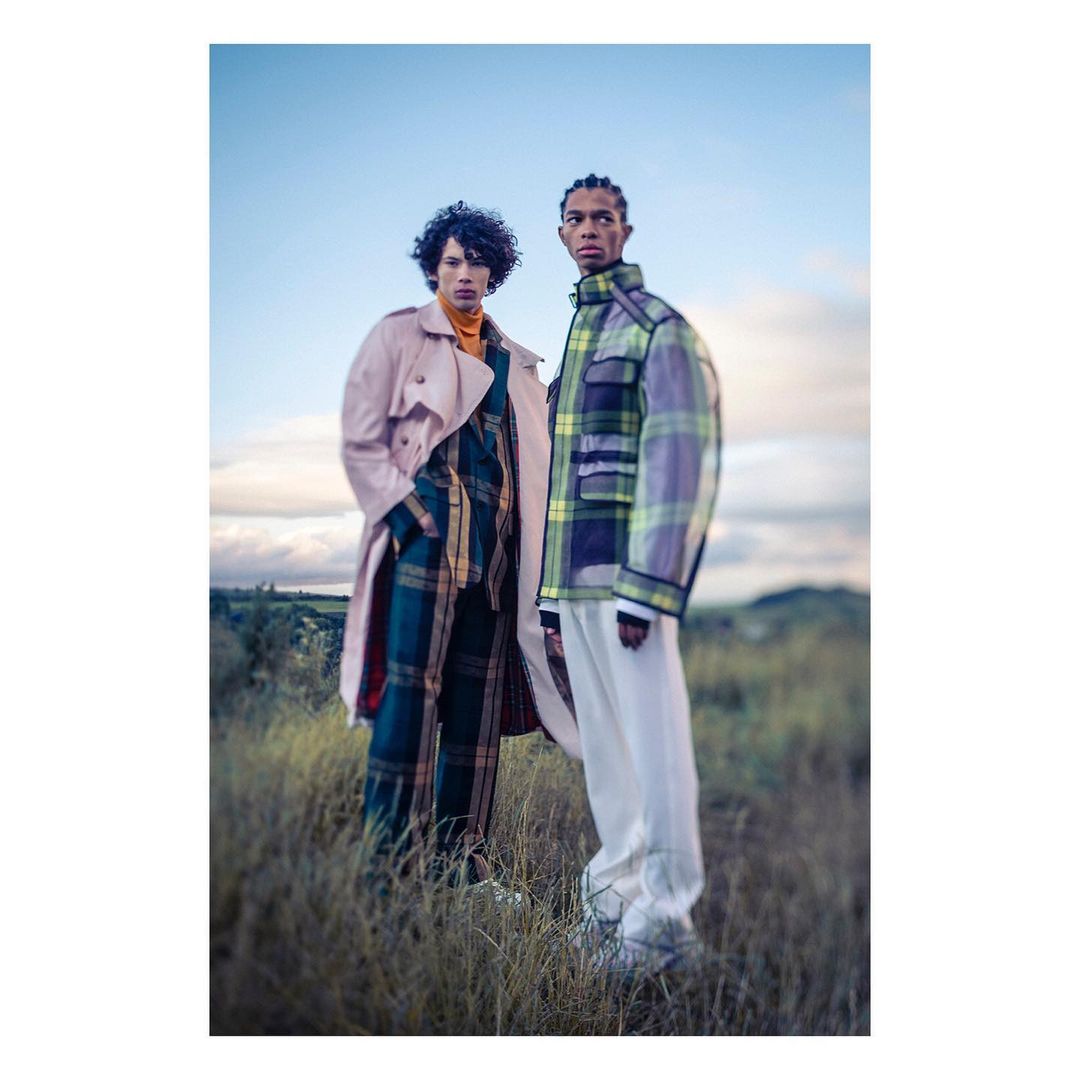 Two fashion models in an outdoor setting showcasing modern apparel, including plaid patterns, a trench coat, and relaxed fit trousers under a blue sky.