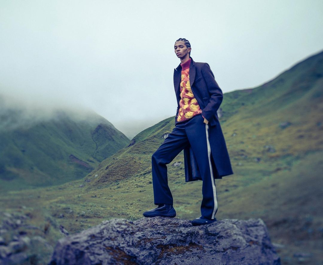 A single adult stands on a rock in outdoor fashion editorial in mountains; fog, green slopes, sweater, wool coat, pants, and shoes visible.